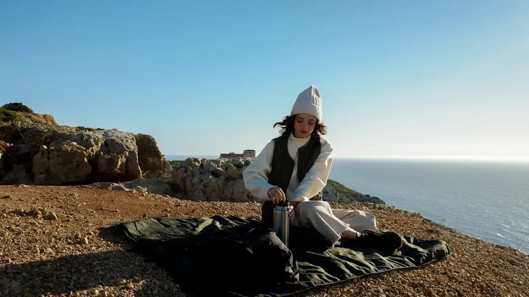 Young woman with thermos enjoys picnic by the ocean on a sunny day.